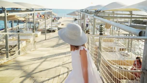 Een klein meisje blond in een witte jurk en een brede hoed loopt op de pier — Stockvideo