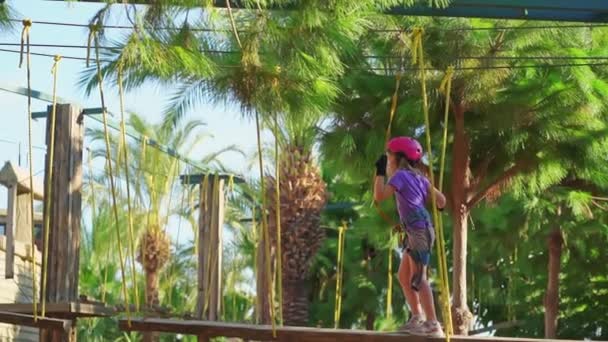 Frightened but brave kid girl in helmet, T-shirt and shorts climbs in rope park — Stockvideo