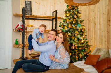 Baba küçük oğlunu elinde tutuyor ve anne de Noel ağacının yanındaki yatağın yanında oturuyor.