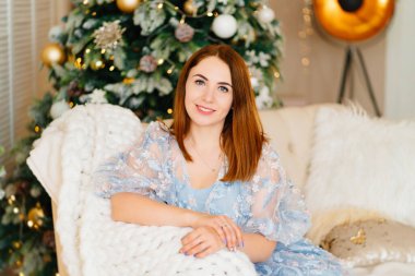 Noel ağacının yanındaki kanepede mavi elbiseli çekici genç bir kadın.