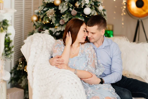 An attractive couple in love in blue clothes on the couch by the Christmas tree. — Fotografia de Stock