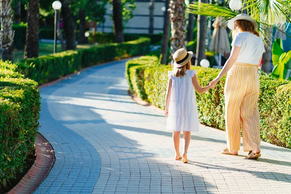 Şapkalı anne ve kızı otelin tropikal parkında yürüyorlar. Arkadan görünüm