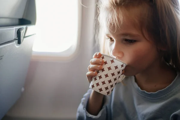En liten blond flicka dricker en drink från en pappersmugg i en flygplansstol. — Stockfoto