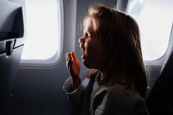 Liten flicka sitter och gäspar i flygplanssitsen vid fönstret. långdistansflygning — Stockfoto