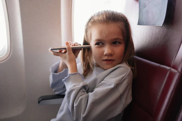 En liten flicka pratar handsfree samtal på telefonen i ett flygplan säte — Stockfoto