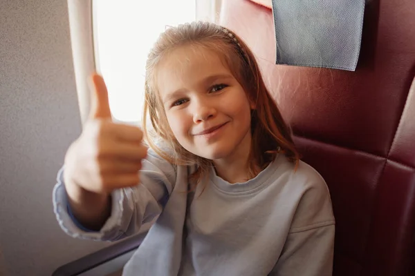 Une drôle de petite fille montre la classe avec le pouce dans un siège d'avion près de la fenêtre — Photo