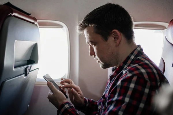 Mann im karierten Hemd SMS am Telefon im Sitz des Passagiers an Bord des Flugzeugs. — Stockfoto