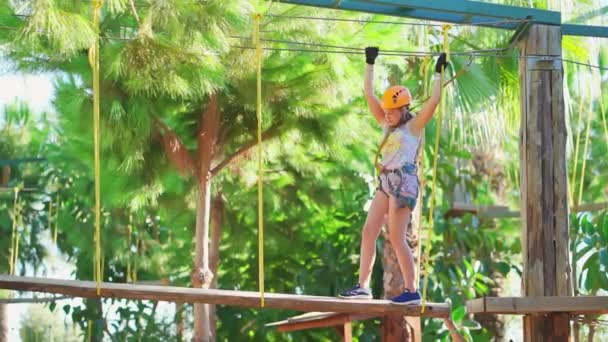 Een vrolijk tiener meisje in een helm, T-shirt en korte broek klimt in een touw park — Stockvideo