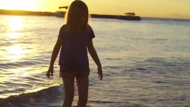 Al atardecer la niña camina a lo largo de la orilla del mar y arroja piedras al agua. — Vídeos de Stock