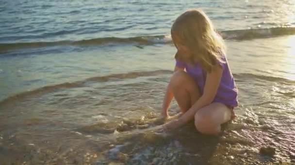 Uma menina bonita senta-se na praia, brincando na areia e com seixos. — Vídeo de Stock