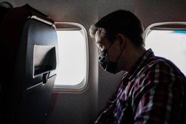 Uçağın penceresinin yanında siyah koruyucu maskeli ve ekose gömlekli bir adam var..