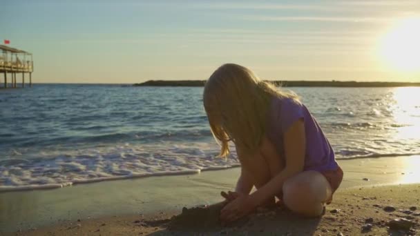 Una linda niña se sienta en la orilla del mar, jugando en la arena y con guijarros. — Vídeos de Stock