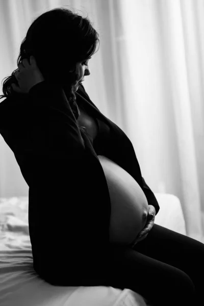 En gravid kvinna i svart jacka med bara bröst och en mage på sängen — Stockfoto