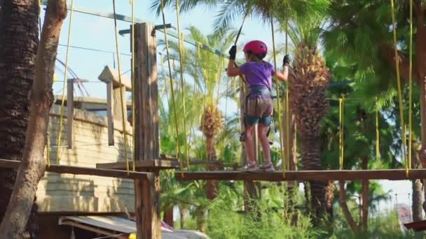 Little girl in helmet, purple T-shirt and shorts afraid and climbs in rope park — Stockvideo