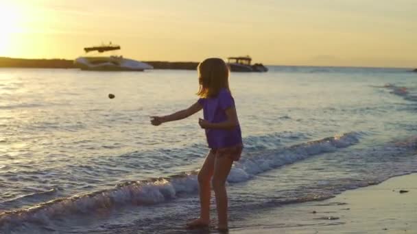 Niña camina a lo largo de la orilla del mar, recoge piedras y las arroja al agua. — Vídeos de Stock