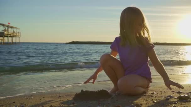 Sevimli küçük bir kız kumsalda oturuyor, kumsalda ve çakıl taşlarıyla oynuyor.. — Stok video