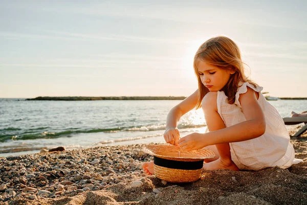 Meisje in witte jurk zit op de kust en verzamelt kiezels en twijgen in stro hoed — Stockfoto