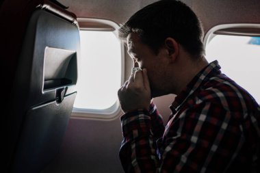 Adam uçaktaki pencerenin yanındaki siyah koruyucu maskeyi ve ekose gömleği düzeltiyor..