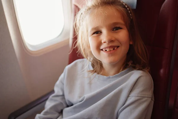 A funny little girl sits in an airplane seat by the window. — Stockfoto