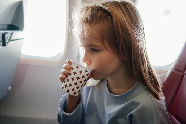 A little blonde girl drinks a drink from a paper cup in an airplane seat. — 图库照片