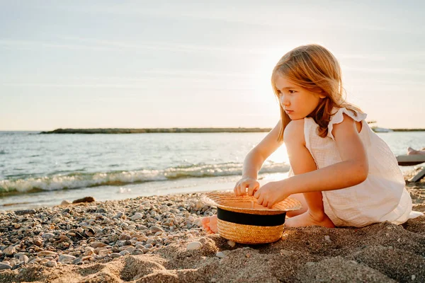 Девушка в белом платье сидит на берегу моря и собирает камешки и веточки в соломенной шляпе — стоковое фото