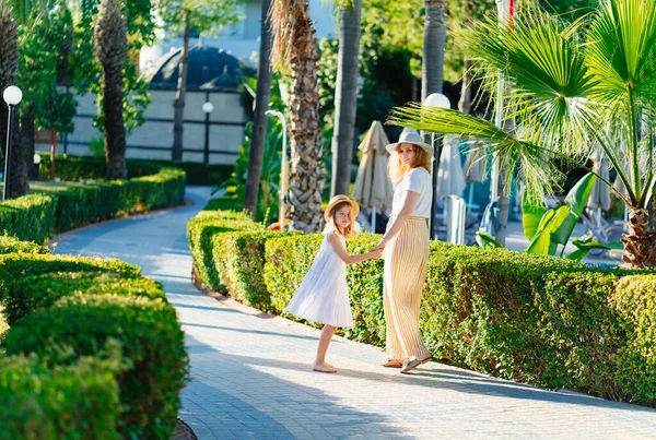 Мама и дочь в шляпах прогуливаются в тропическом парке отеля. view from back — стоковое фото