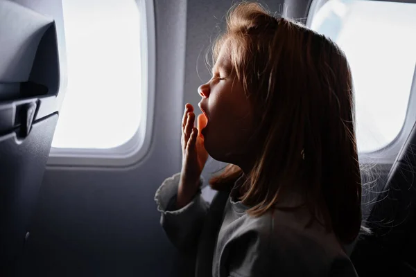 Little girl sits and yawns in airplane seat by window. tedious long flight — Stockfoto