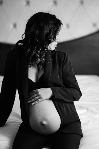 En gravid kvinna i svart jacka med bara bröst och en mage på sängen — Stockfoto