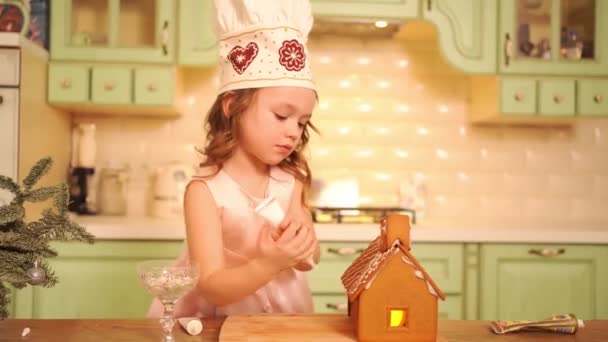 Şefin şapkası takmış küçük bir kız zencefilli kurabiye evini şekerle süslüyor.. — Stok video