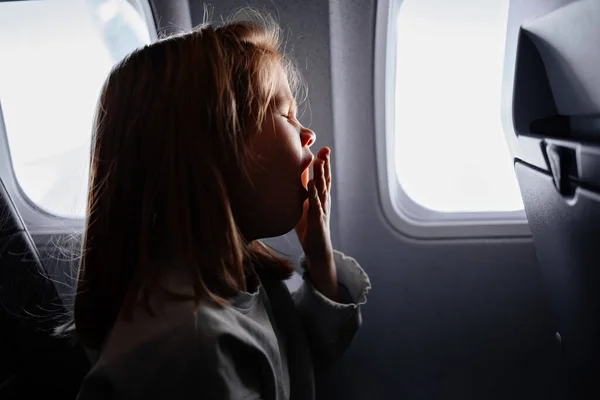 Little girl sits and yawns in airplane seat by window. tedious long flight — Stockfoto