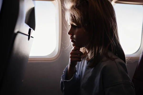 Una niña triste se sienta en un asiento de avión junto a la ventana. — Foto de Stock