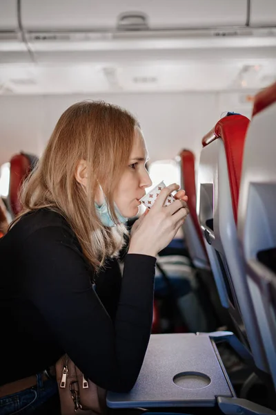 Una mujer con una máscara protectora bebe una bebida de una taza de papel a bordo del avión. — Foto de Stock