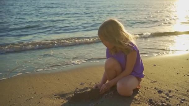 Una linda niña se sienta en la orilla del mar, jugando en la arena y con guijarros. — Vídeos de Stock