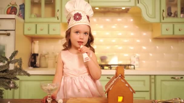 Una niña con gorra de chefs decora una casa de pan de jengibre con glaseado de azúcar. — Vídeos de Stock