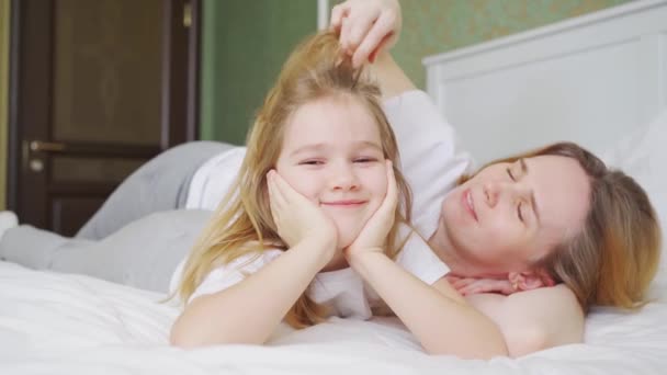 Mamá y su hija están acostadas en la cama. mamá ajusta el pelo de sus hijas. — Vídeos de Stock