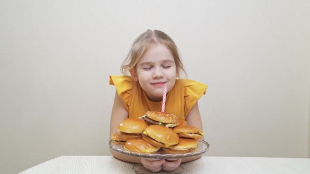 O fetiță amuzantă face o dorință și suflă o lumânare pe un tort de hamburger. — Videoclip de stoc