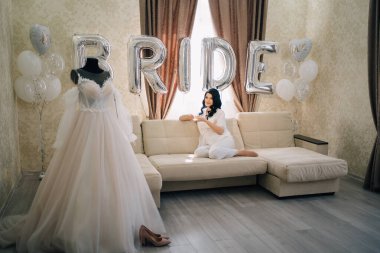 Gelin beyaz önlüklü bir odada balonlarla kanepede çay içiyor.