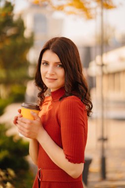 Kahverengi elbiseli esmer bir kadın sonbahar parkında kahve içiyor..