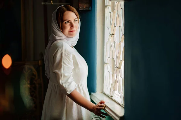 Una mujer con pañuelo en la cabeza y túnicas de colores claros por la ventana en la iglesia. — Foto de Stock