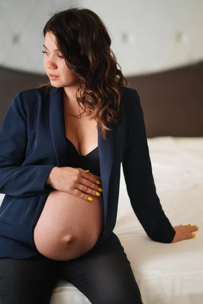 En gravid kvinna i svart jacka med bara bröst och en mage på sängen — Stockfoto