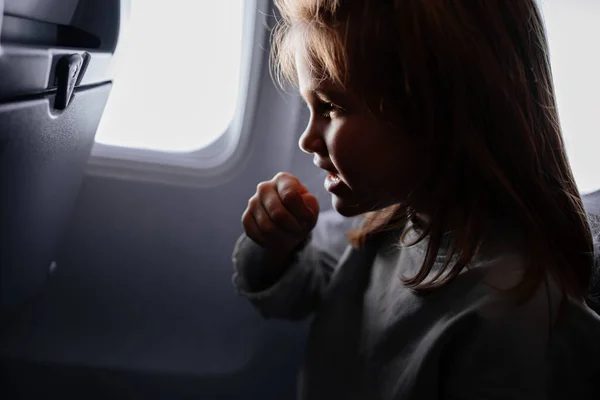 En liten flicka sitter och hostar i ett flygplanssäte vid fönstret. — Stockfoto