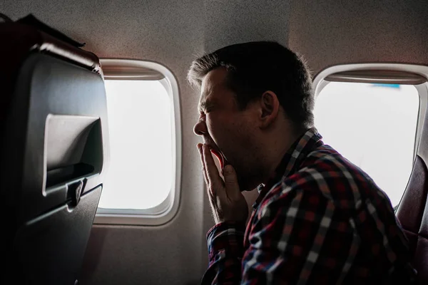 Un hombre con una camisa a cuadros se sienta junto a la ventana del avión y bosteza. vuelo largo. — Foto de Stock