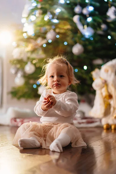 Uma menina bonita em roupas leves senta-se sob uma árvore de Natal. — Fotografia de Stock