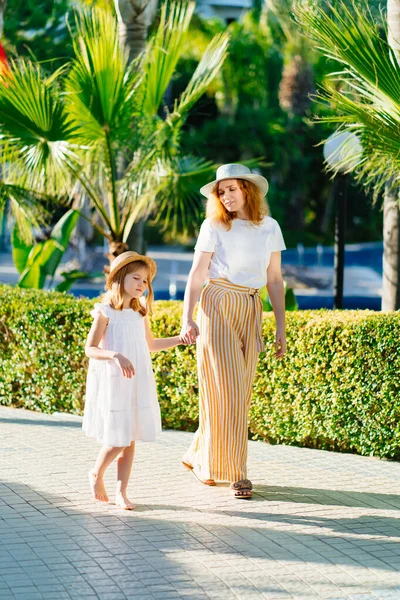 Şapkalı anne ve kızı otelin tropikal parkında yürüyorlar.