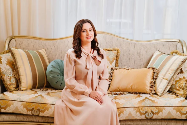 Una mujer morena en un elegante vestido beige se sienta en un sofá retro clásico. — Foto de Stock