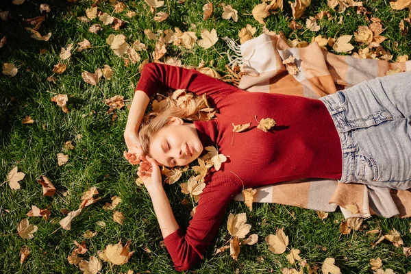 Atraktivní blondýnka leží na zeleném trávníku s podzimními listy. Stock Snímky