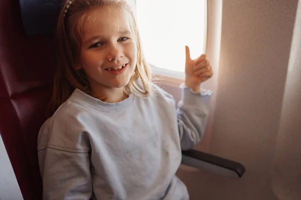 A funny little girl shows the class with thumb in an airplane seat by the window — Stockfoto