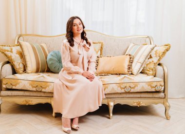 Zarif bej elbiseli esmer bir kadın klasik bir retro kanepeye oturur..