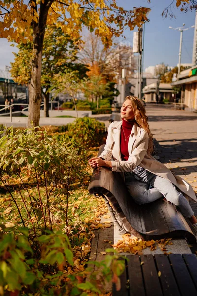 Palto giymiş güzel bir sarışın ve sonbahar parkında bankta kot pantolon.. — Stok fotoğraf