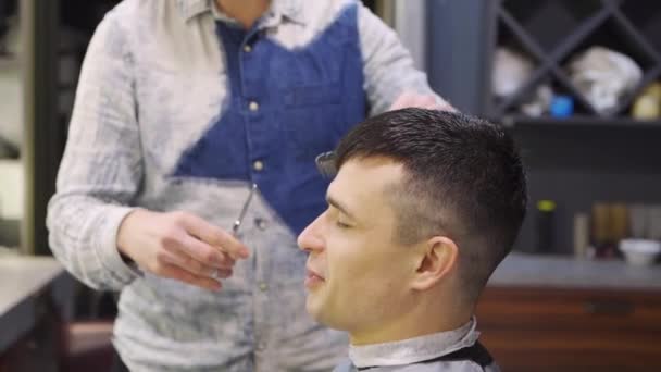 Cabeleireiro faz um corte de cabelo para um homem morena com uma tesoura em uma barbearia — Vídeo de Stock